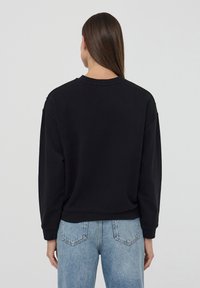 Felpa nera con colletto tondo e spalle scese, caratterizzata da una vestibilità comoda e polsini a coste. Abbinata a jeans azzurri chiari.
