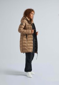 Tans kleurige pufferjack met een capuchon, voorzien van een gestikt ontwerp, zijzakken en een zwarte rits. Gecombineerd met zwarte broek en witte sneakers.