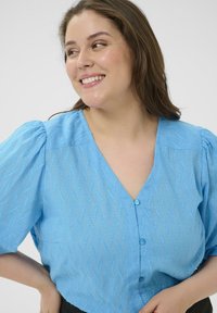 Blouse bleu clair avec manches bouffantes, col en V et fermeture à boutons ; présente un motif ondulé texturé sur toute la surface.