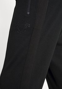 Černé sportovní kalhoty s hladkou texturou a bočním pruhem. Obsahují lesklé kamínky tvořící logo na oblasti boků.