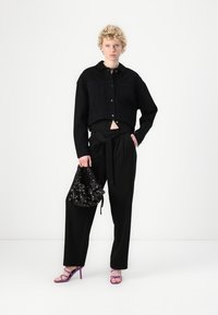Black cropped denim jacket with button closure, high-waisted black trousers with a tie, purple high-heel sandals, and a patterned black handbag.