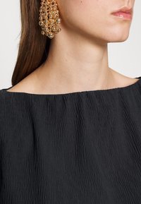 Femme portant de grandes boucles d’oreilles pendantes en perles dorées et un haut noir texturé décolleté, vue du cou au menton.