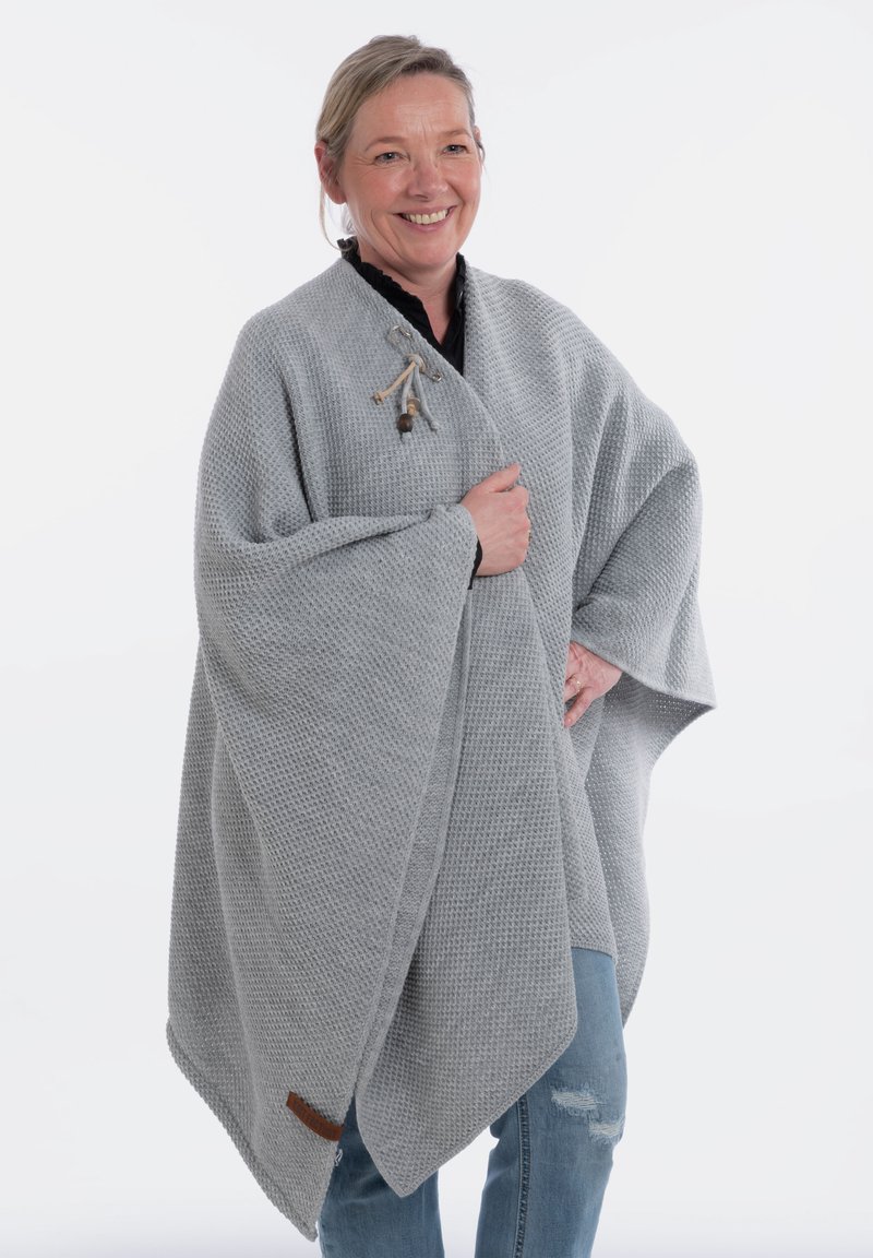 Poncho texturé gris clair avec un motif tissé, doté d'une fermeture à bascule en bois, tombant sur un jean décontracté.