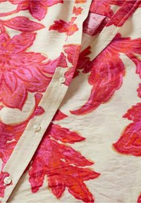 Camicia a maniche corte con audaci motivi floreali rosa su sfondo crema, caratterizzata da una chiusura con bottoni e un tessuto morbido e texturizzato.