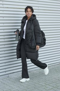 Melnā pūderveida jaka ar kapuci, divām sānu kabatām un oversized piegriezumu, valkāta ar melnām biksēm un baltām sporta kurpēm, turot ūdens pudeli.