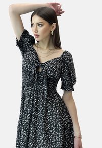 Vestido negro con estampado floral blanco, mangas abullonadas y escote con lazo. Tejido suave con un corte relajado y cintura fruncida. Se llevan collares y pendientes en oro con capas.