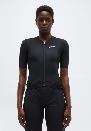 Μαύρο κοντομάνικο ποδηλατικό τζέρσεϊ με φερμουάρ και λογότυπο "poc", φορεμένο από ένα άτομο που στέκεται μπροστά σε απλό ανοιχτό φόντο.