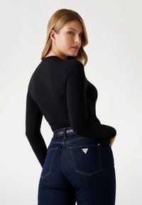 Top nero a maniche lunghe abbinato a jeans blu scuro. I jeans presentano un logo triangolare sulla tasca posteriore e una cintura in pelle liscia.