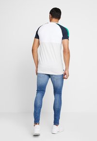 Camiseta de manga corta en bloques de color con secciones blanca, azul marino y verde; combinada con jeans ajustados azul y zapatillas blancas.