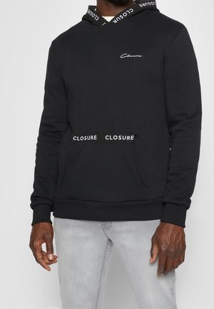 Homme portant un sweat à capuche noir avec "Closure" imprimé sur les cordons de la capuche et la poche avant, associé à un jean gris clair.