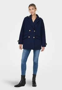 Manteau bleu marine à double boutonnage avec des boutons dorés, poches latérales et épaules structurées, associé à un jean bleu et des bottines noires.