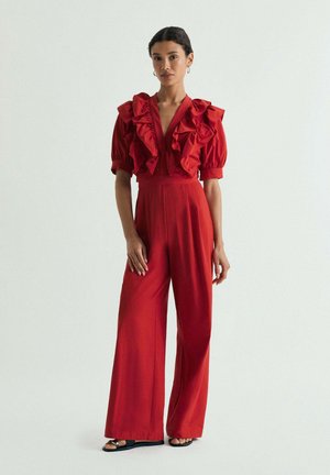 Mujer de pie que lleva un mono rojo con mangas cortas abullonadas, grandes volantes en el pecho, piernas anchas y zapatos negros abiertos en la punta.