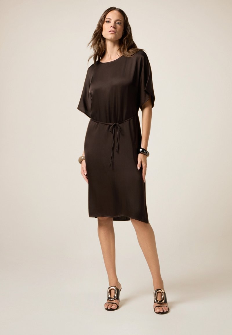 Mujer vestida con un vestido marrón oscuro a la altura de la rodilla, con mangas con volante, cintura ceñida, tacones con grandes hebillas metálicas y pulseras a juego, de pie.
