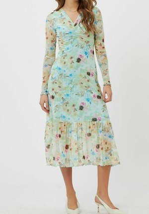 Femme portant une robe midi à manches longues, col en V, cintrée avec un motif floral bleu clair, ourlet à volants et des talons slingbacks blancs, debout devant un fond blanc.
