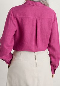 Camicia in lino fucsia con colletto a bottoni, dettaglio di piega posteriore e maniche lunghe, abbinata a una gonna beige con zip invisibile.