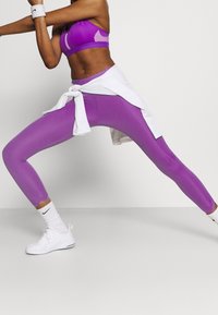 Leggings viola realizzati in materiale elastico, con un design aderente e una texture liscia, abbinati a una maglietta bianca annodata in vita.