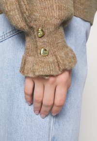 Maglione marrone lavorato a maglia con maniche a volant, con due bottoni dorati a forma di cuore vicino al polsino, indossato sopra un paio di jeans blu.
