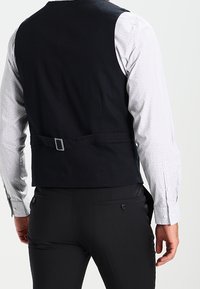 Schwarze Stoffweste mit einem zusammenziehbaren Riemen und einer Metallschnalle, getragen über einem hellen, gemusterten Hemd, kombiniert mit dunklen, maßgeschneiderten Hosen.