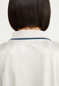 Nike pikétröja i ljusbeige med vertikal, subtil branding. Har en kontrasterande mörk krage och en liten logotyp nära halsringningen.