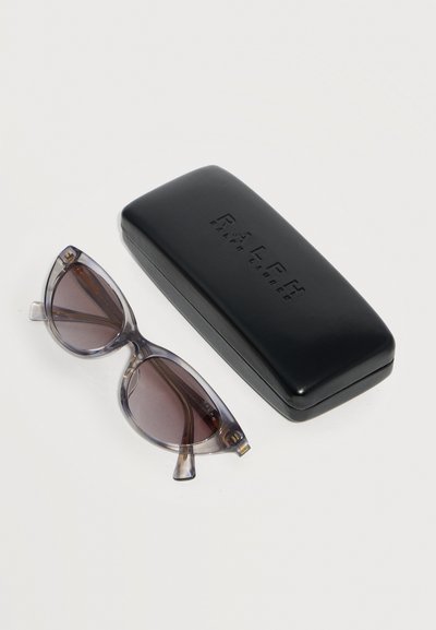 Ημιδιαφανή γκρι γυαλιά ηλίου cat-eye με σκούρους φακούς δίπλα σε κλειστή μαύρη σκληρή θήκη με το λογότυπο Ralph Lauren σε λευκό φόντο.