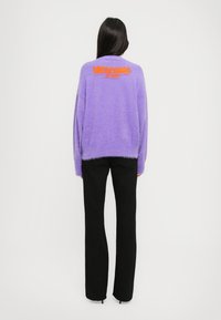 Fioletowy puszysty sweter z pomarańczowym napisem "MOSCHINO" na plecach, w zestawieniu z dopasowanymi czarnymi spodniami. Widoczna faktura i luźny krój.