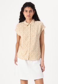 Chemise beige à manches courtes avec un motif de coquillage, dotée d'un col boutonné et de boutons blancs, associée à une jupe en denim blanche.
