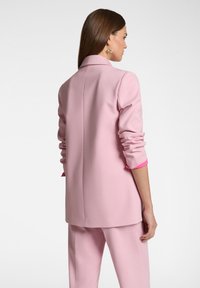 Blazer rosa chiaro con maniche lunghe, colletto a revers e polsini interni rosa a contrasto, che mette in mostra una vestibilità sartoriale e una texture liscia.