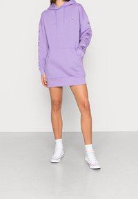 Lavendel hoodie-kjole med lange ærmer, frontlomme og logodetalje. Afsluttet med ribbede manchetter og kant. Båret med hvide sneakers.