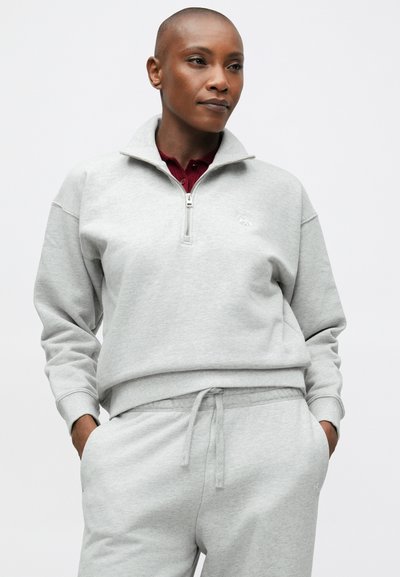 Osoba s krátkymi vlasmi, oblečená v svetlošedých zip-up mikine a zodpovedajúcich teplákoch, stojí pred jednoduchým bielym pozadím.