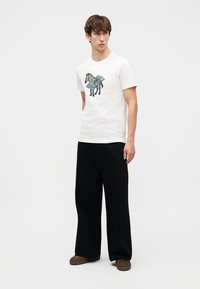 Νεαρός άνδρας που φοράει λευκό μπλουζάκι με γραφικό ζέβρα και κύκλωμα, μαύρο φαρδύ παντελόνι και καφέ παπούτσια, στέκεται μπροστά σε λευκό φόντο.