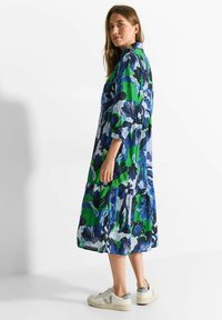 Langes, gestuftes Kleid mit einem lebhaften Blumenmuster in Blau und Grün, mit Kragen und Dreiviertelärmeln, kombiniert mit neutralen Sneakers.