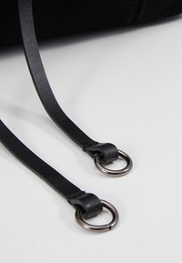 Dvě černé kožené popruhy s metalickými kroužky připevněnými k nim, vystavené na bílé ploše, s částí černého předmětu v pozadí.