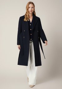 Granatowy płaszcz dwurzędowy z dużymi guzikami, szerokimi klapami i paskiem w talii, zestawiony z białymi spodniami i granatowym swetrem. Miękka tekstura.