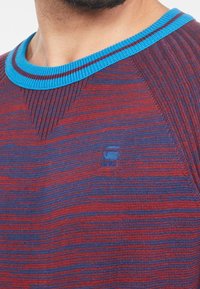 Pull en tricot à rayures rouges et bleues. Présente un col rond côtelé avec un accent bleu. Petit logo sur la poitrine.
