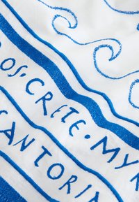 Tissu blanc avec des volutes décoratives brodées en bleu et un texte brodé en majuscules partiellement visible.