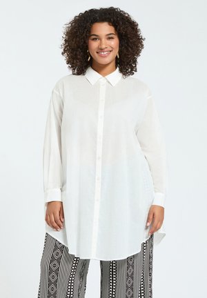 Lächelnde Frau mit lockigem Haar trägt ein langes weißes Hemd mit Knöpfen und schwarz-weiß gemusterte weite Hosen, steht vor einem schlichten Hintergrund.