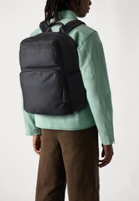 Sac à dos noir avec plusieurs poches zippées, porté sur une veste vert clair. Il dispose de sangles ajustables et d'une finition lisse et mate.