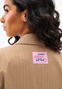 Žena s tmavými kučeravými vlasmi oblečená v béžovom blejzri s tenkými prúžkami, na ktorom je ružová náplasť s nápisom „ROMANCE“ a rukou písaným textom na chrbte.