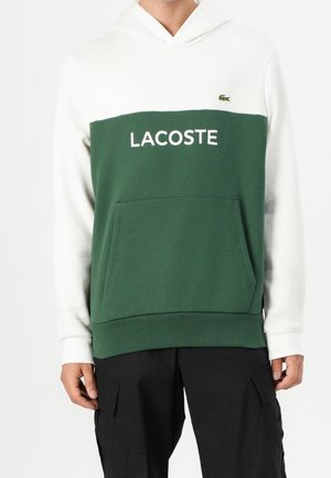 Sudadera con capucha de parte superior y mangas blancas, pecho y bolsillo verdes, texto "LACOSTE" y pequeño logo verde de cocodrilo en el lado izquierdo del pecho.