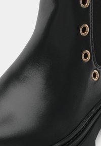 Botín negro de cuero con acabado brillante, puntera redonda y detalles de ojales dorados. Presenta una textura suave y una suela resistente.