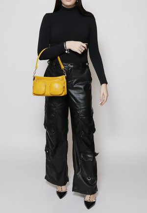 Persona con dolcevita nero e pantaloni cargo in pelle nera a gamba larga, che tiene una borsa gialla e indossa tacchi neri a punta.