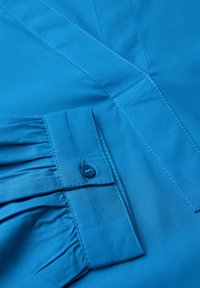 Blauwe shirt met een geplooide mouwmanchet en een enkele knoopsluiting. Glad materiaal met subtiele stikseldetails.