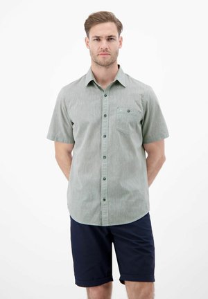 Mężczyzna w jasnozielonej koszuli z krótkim rękawem na guziki i ciemnoniebieskich szortach stojący na białym tle.