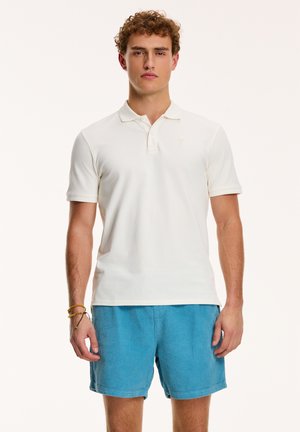 Camisa polo blanca con un cuello corto y tres botones, combinada con pantalones cortos azules de textura. El conjunto presenta un sutil detalle de logo en el pecho.