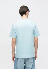 Νεαρό άτομο με κοντά σκούρα μαλλιά που φοράει ανοιχτό μπλε t-shirt και δύο τόνων ανοιχτό μπλε τζιν, στέκεται στραμμένο σε έναν απλό λευκό τοίχο.