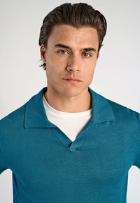 Niebieska dzianinowa koszulka polo z dekoltem w serek, krótkimi rękawami i teksturowanym wykończeniem. Nosi się ją na białym t-shircie z okrągłym dekoltem, na jednolitym tle.