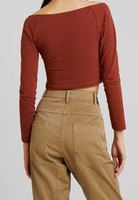 Rostfarbener, langärmeliger Crop-Top mit weitem Ausschnitt; kombiniert mit khakifarbenen Hosen, die zwei Taschen auf der Rückseite und Nahtdetails aufweisen.