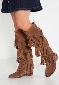 Botas de ante marrón hasta la rodilla con detalles de flecos en capas, punta afilada y tacón bajo apilado, colocadas sobre un fondo neutro.