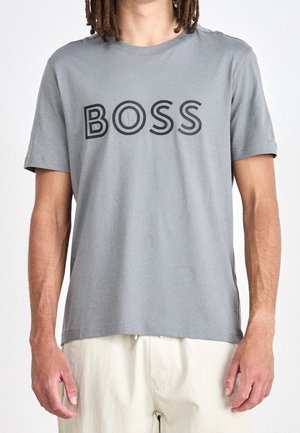 Person trägt ein graues Kurzarm-T-Shirt mit dem Wort "BOSS" in großen schwarzen Buchstaben auf der Brust und hellfarbene Hose.