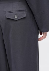 Κοντινή λήψη γκρίζων παντελονιών με τσέπη στο πίσω μέρος που κλείνει με κουμπί και μέρος ενός χεριού που ακουμπάει δίπλα στο σώμα, σε λευκό φόντο.